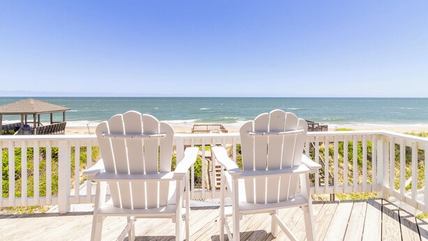 A wooden deck with white Adirondack chairs facing a beach. - Sun Realty stunning oceanfront Nags Head vacation home with elevator! (Nags Head)