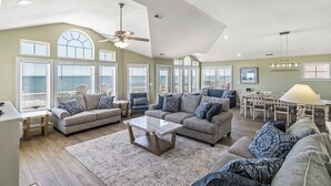 A spacious living room with multiple seating areas, large windows, and a ceiling fan. - Sun Realty stunning oceanfront Nags Head vacation home with elevator! (Nags Head)