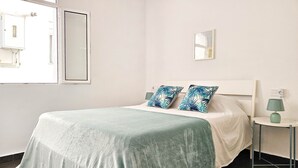 Un lit bien fait, recouvert d’une couvre-lit verte et agrémenté de deux coussins décoratifs, une table de chevet avec une lampe, et un miroir fixé au mur.