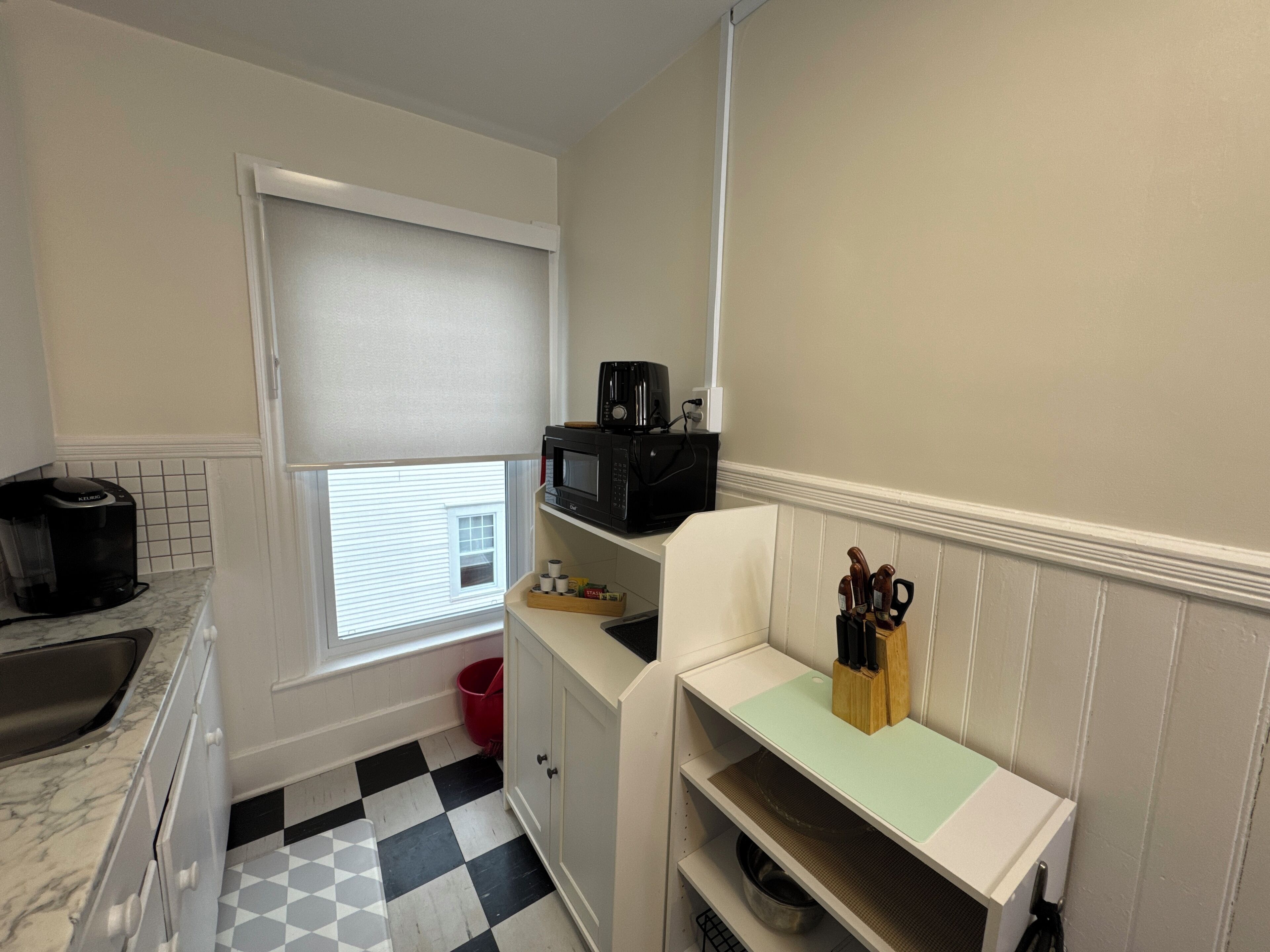 A compact kitchen with a checkered floor, a coffee maker, a microwave, and a small window.