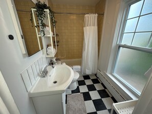 Baño con lavamanos, inodoro y zona de ducha blancos. Cuenta con una ventana que proporciona luz natural y vistas al exterior.