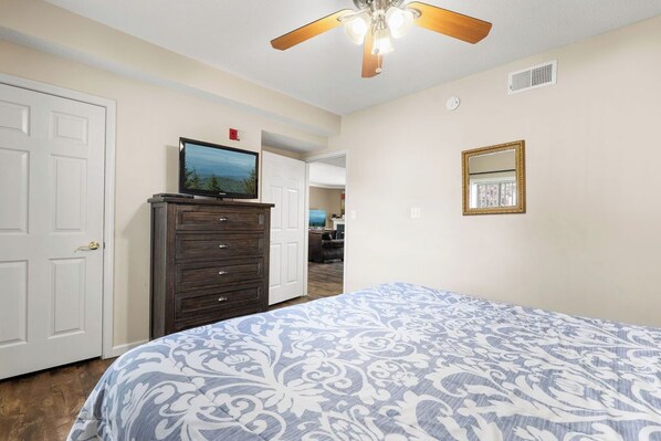 A bedroom with a bed, dresser, television, ceiling fan, and a door leading to another room.