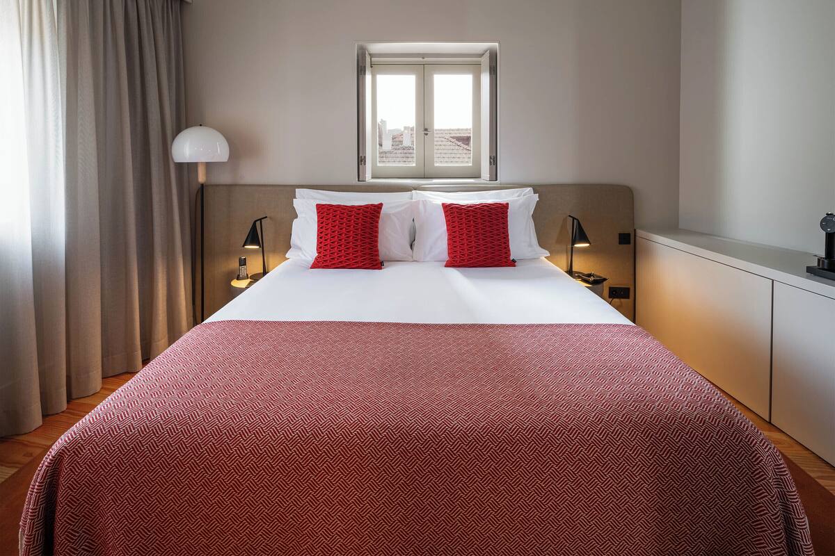 A hotel room with a large bed, two bedside lamps, a window, and a view of the cityscape.