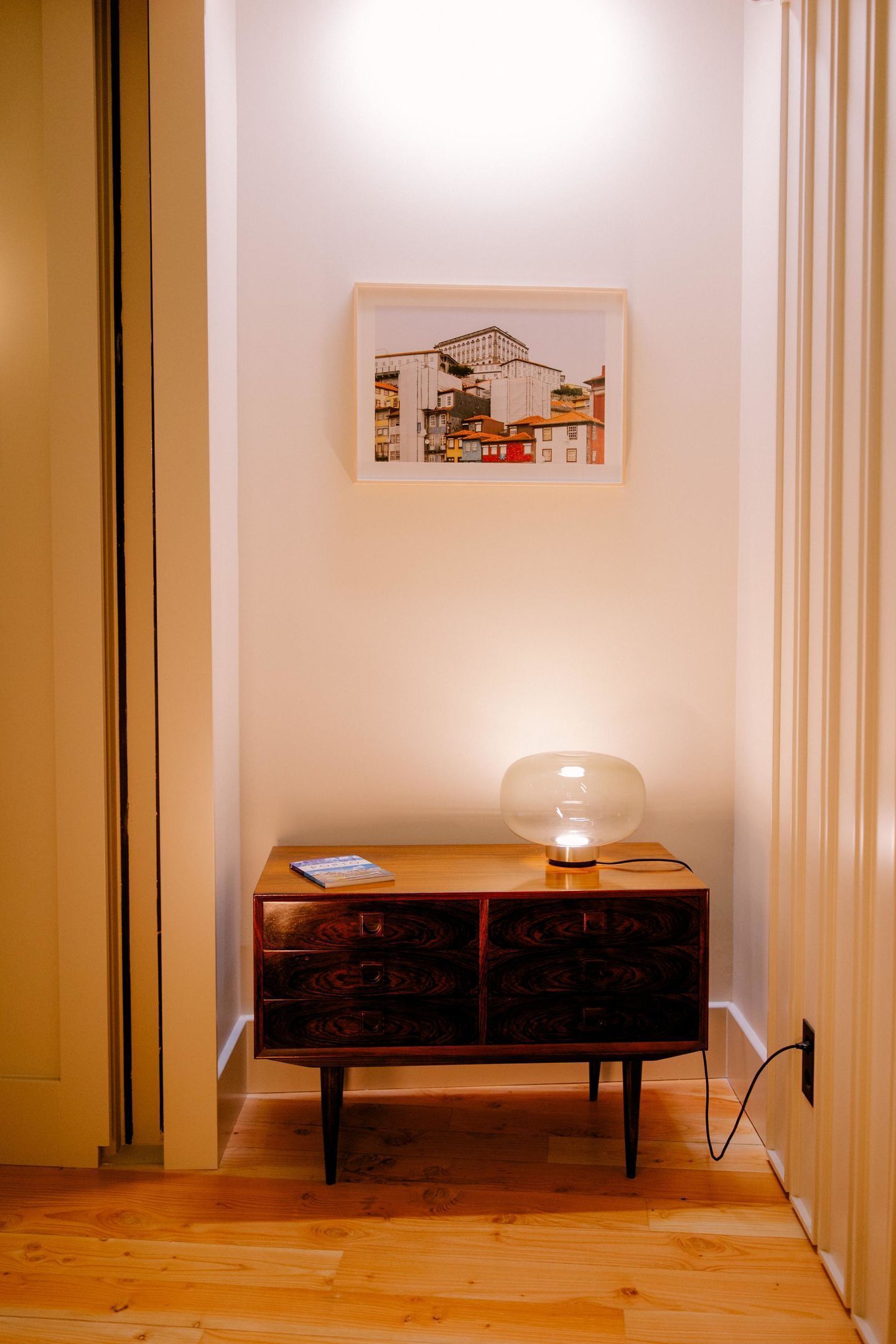 Un couloir doté d’un plancher en bois, une commode sombre en bois avec des tiroirs, un tableau encadré accroché au mur et une lampe de table posée sur la commode.