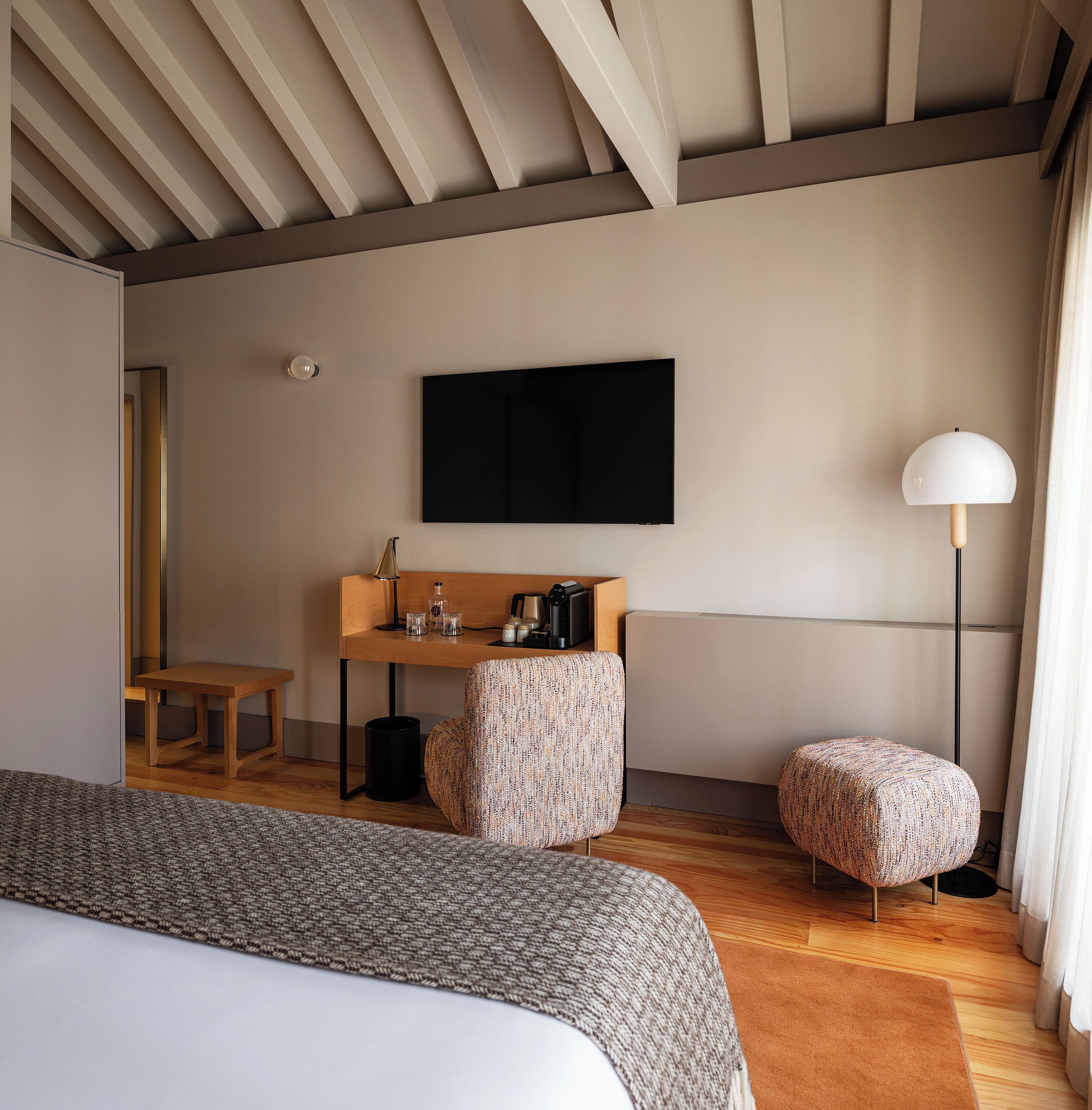 A modern hotel room with a large bed, a desk with a lamp, a TV, and a small stool.