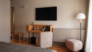 A modern hotel room with a large bed, a desk with a lamp, a TV, and a small stool.