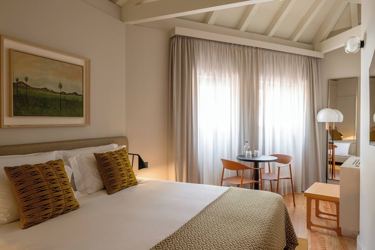 A hotel room with a bed, a desk, and a chair. There is a painting on the wall and a window with curtains.