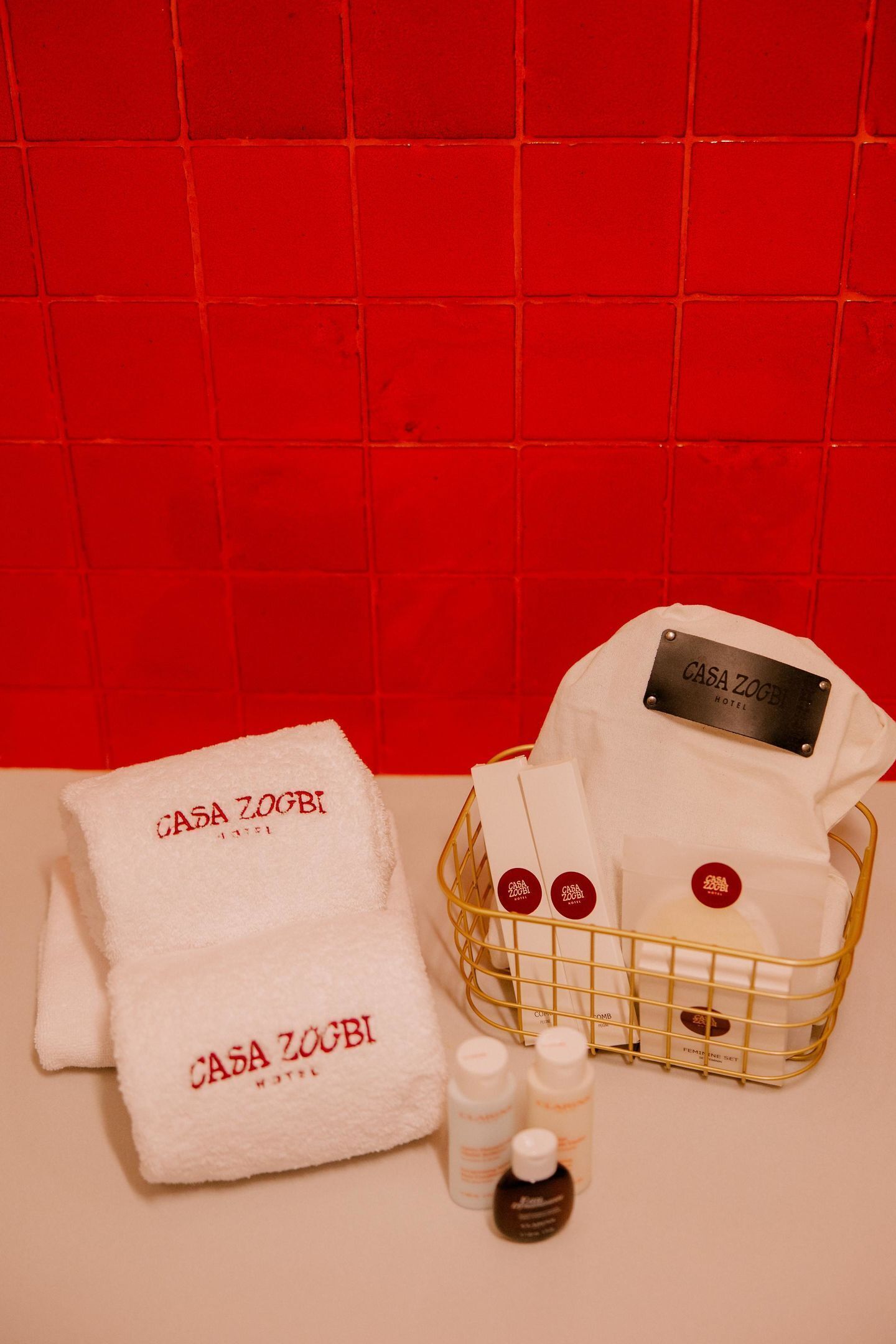 Ein Badezimmer mit roter Fliesenwand, einem weißen Handtuch, einem weißen Bademantel und einem goldenen Korb mit Toilettenartikeln.