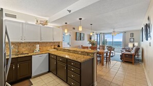 A modern kitchen with dark cabinetry, a granite countertop, and a dishwasher. There is a dining area with a table and chairs, and a balcony with a view. - 🏝️Beachfront-Spacious Balcony-Pool-Romar Place 305🏝️ (Orange Beach)
