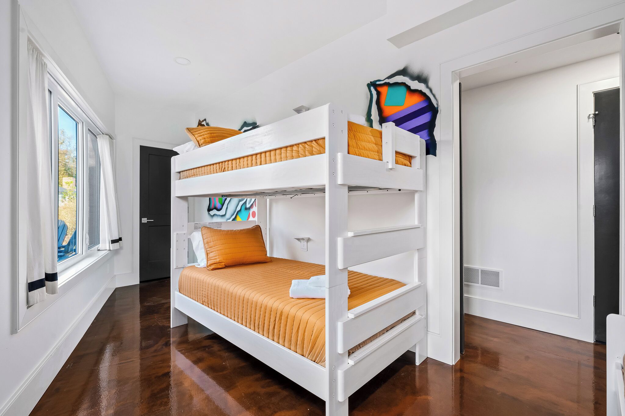 Een slaapkamer met een stapelbed, een raam, een deur en een muur met een kleurrijk schilderij.