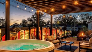 Un jacuzzi bajo una pérgola con luces colgantes, asientos exteriores y una chimenea.