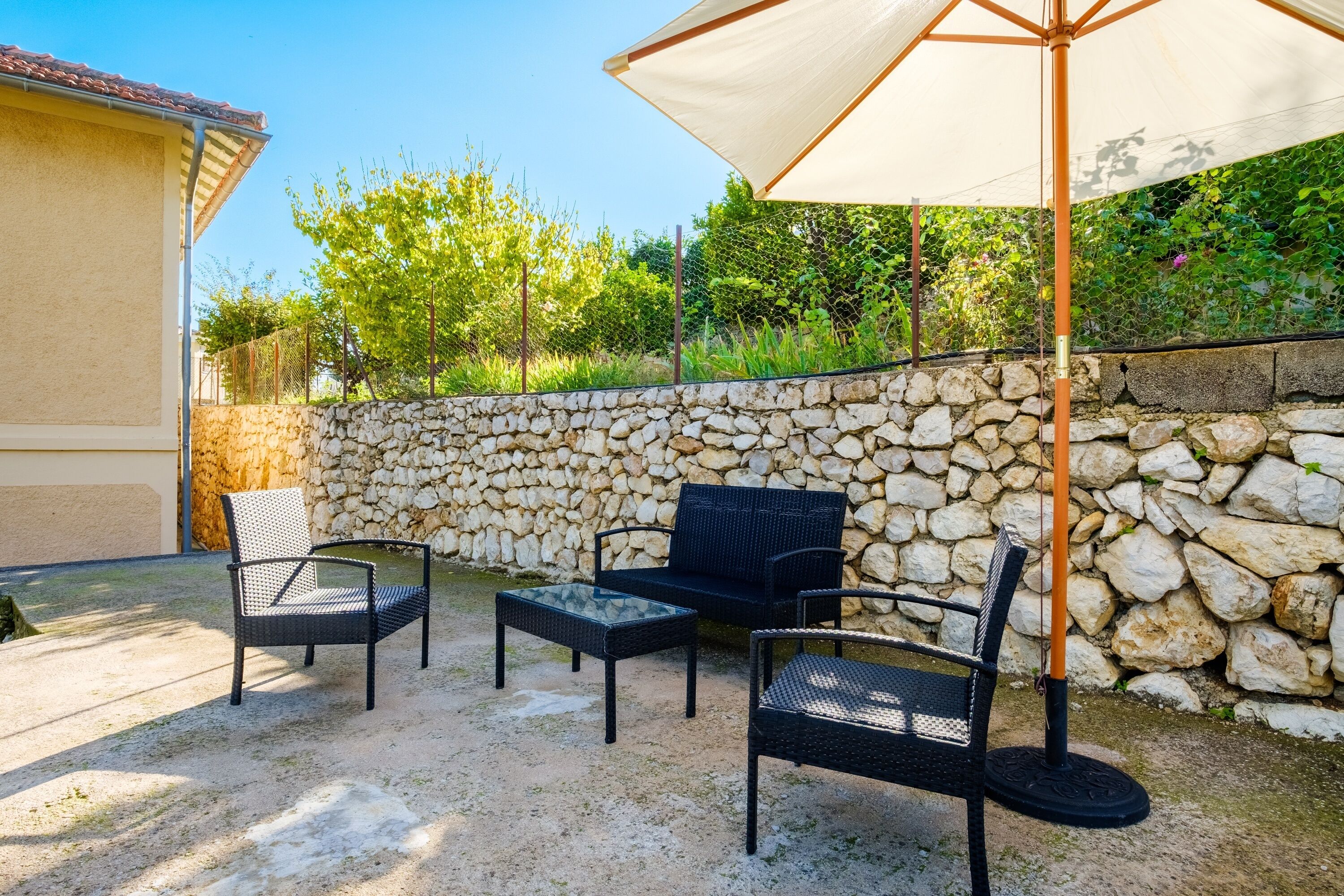 Un espace extérieur aménagé en terrasse, avec des meubles en osier, un mur en pierre et un grand parasol.