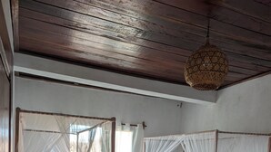 Una habitación con dos camas de madera, cada una con dosel y almohadas amarillas. Hay una mesita con una lámpara y un objeto decorativo entre las camas. Un ventilador está en el suelo, junto a una de las camas.