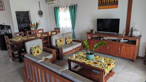 Una sala de estar con un sofá, una mesa de centro con mantel de girasol, un televisor sobre un soporte de madera y un cuadro en la pared.
