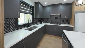A modern kitchen with dark cabinetry, a white countertop, a hexagonal backsplash, and a window with a black roller blind.