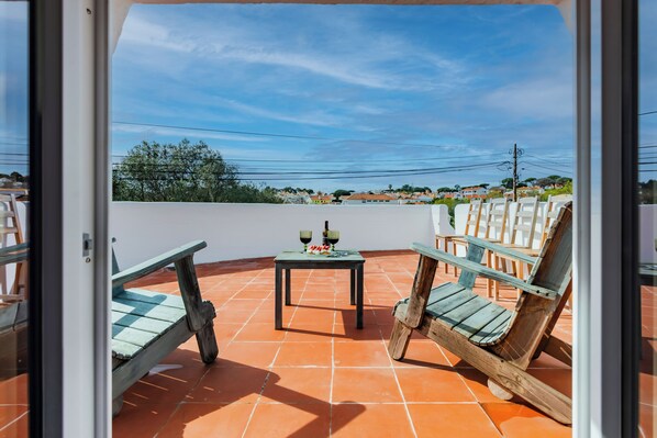 A terrace with a table and two wooden chairs, a tiled floor, and a view of houses in the distance.