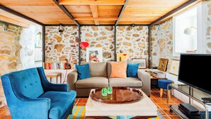 A cozy living room with a stone wall, wooden ceiling beams, a blue armchair, a beige sofa, and a flat-screen TV. - Casalinho de Bicesse (Cascais)