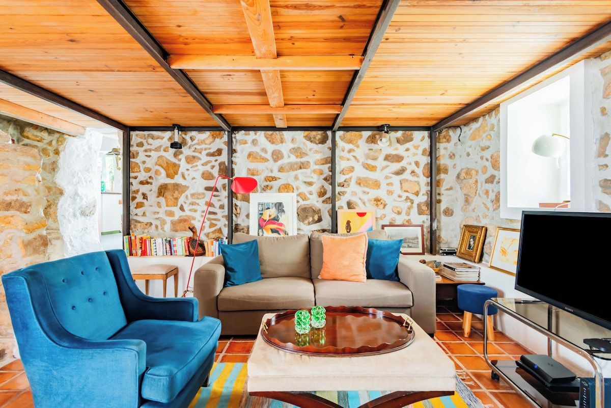 Un salón acogedor con pared de piedra, techo de madera, un sofá, un sillón azul, una mesa de centro y un televisor.
