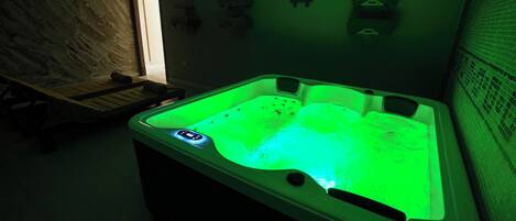 A spa room with a hot tub illuminated by green lighting, a wooden bench, and a tiled wall.