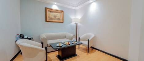 A room with a light blue wall, a framed artwork, a white sofa, two armchairs, a small table with plates, a floor lamp, and a coffee machine.