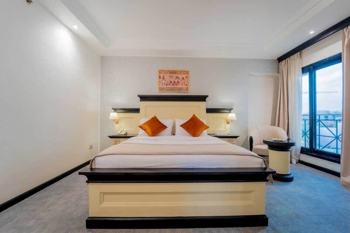 Ein Hotelzimmer mit einem großen Bett, zwei Nachttischen, einem Sessel und Blick auf die Stadt durch das Fenster.