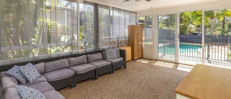 A sunlit room with a sofa, a ceiling fan, and a view of a swimming pool through a sliding glass door.