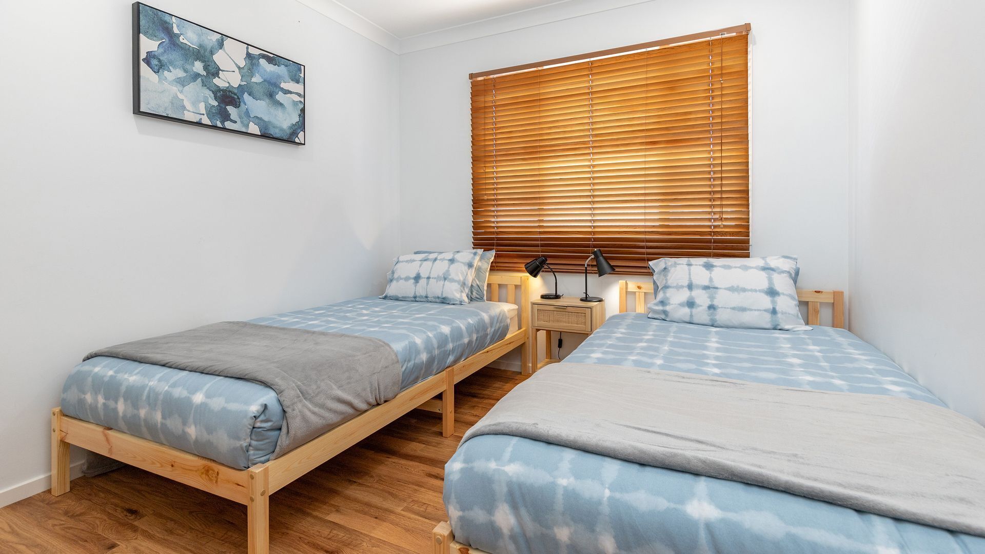 Une chambre avec deux lits simples, des têtes de lit en bois, une table de chevet avec une lampe et une fenêtre avec des stores.