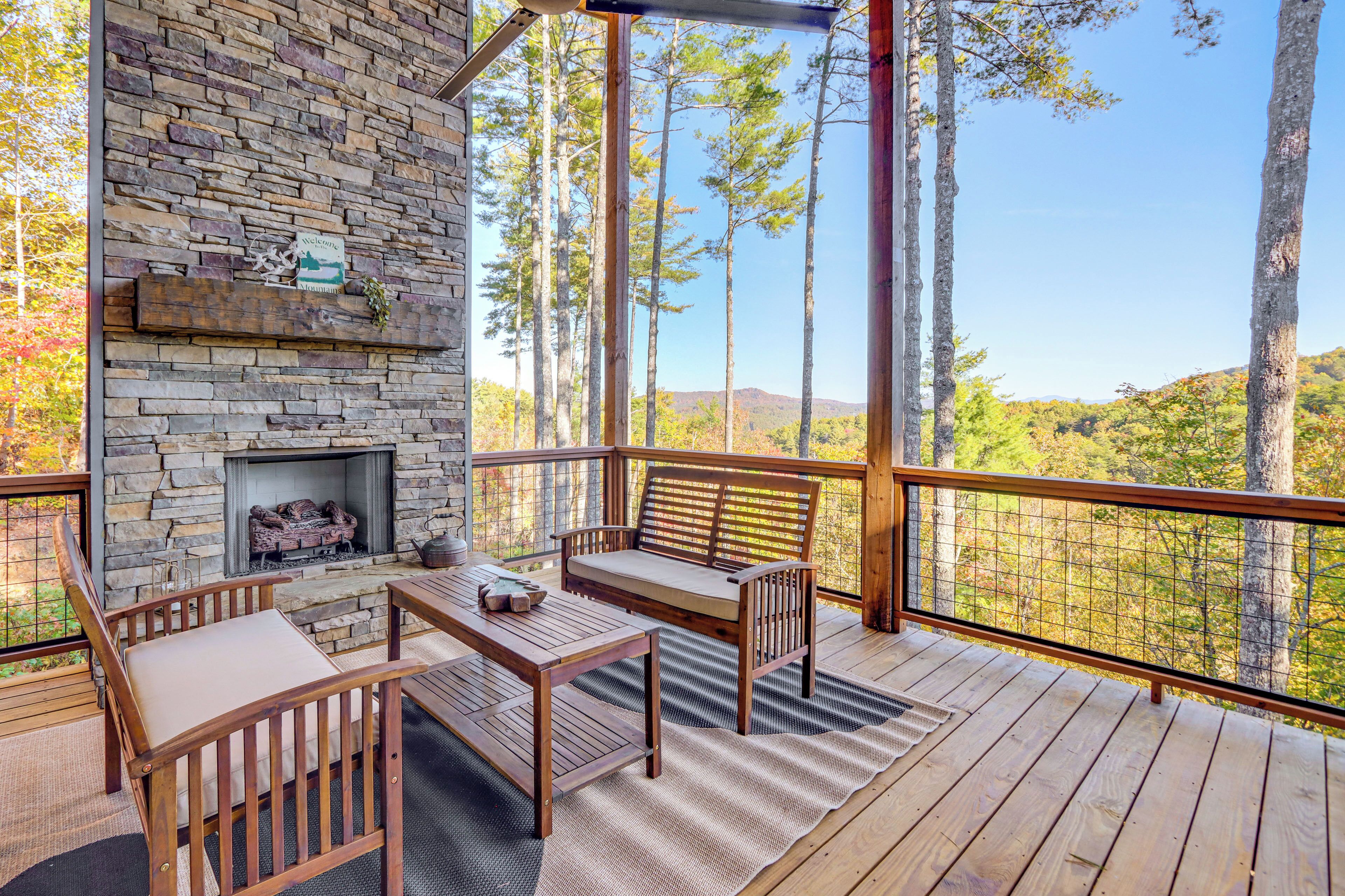 Una terraza de madera con una chimenea de piedra, asientos y vistas a altos árboles y colinas.