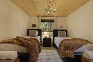 A bedroom with two beds, a nightstand with a lamp, a window with a view of trees, and a wooden ceiling.