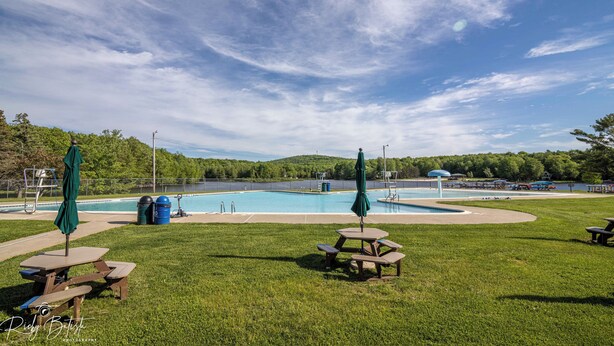 A swimming pool with picnic tables and umbrellas, surrounded by a grassy area and trees.