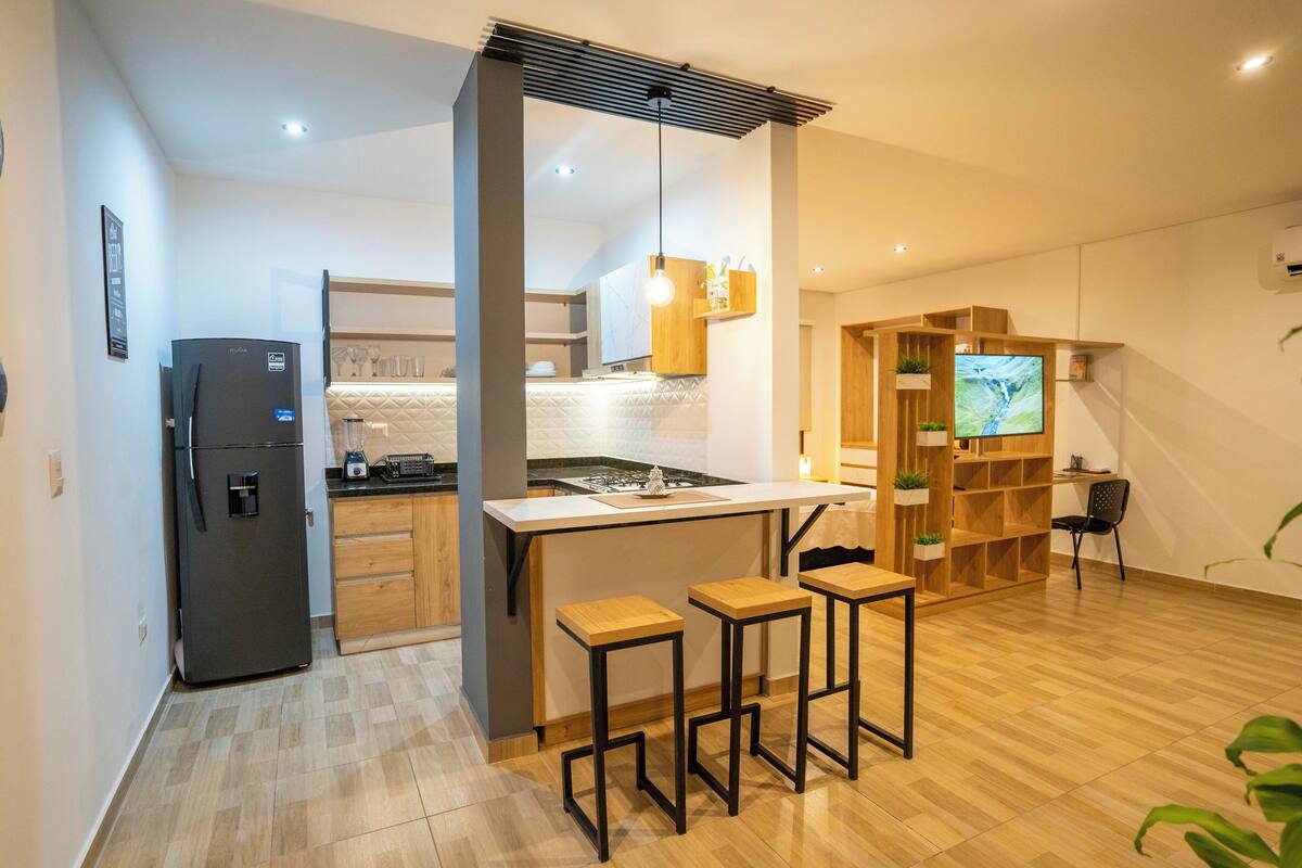 Cozinha compacta com geladeira, área de refeições com banquetas, estante de madeira e televisão.