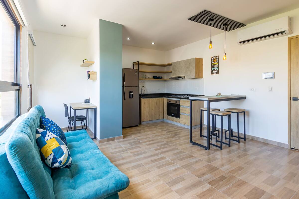 Uma cozinha moderna com ilha central, armários de madeira e forno embutido. Um sofá azul com almofadas estampadas está em primeiro plano.