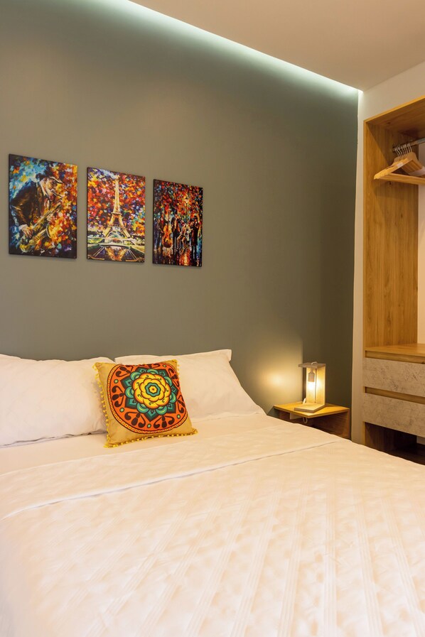 Un lit bien fait, agrémenté d’un coussin aux couleurs vives, une table de chevet avec une lampe, et trois tableaux aux couleurs éclatantes accrochés au mur.