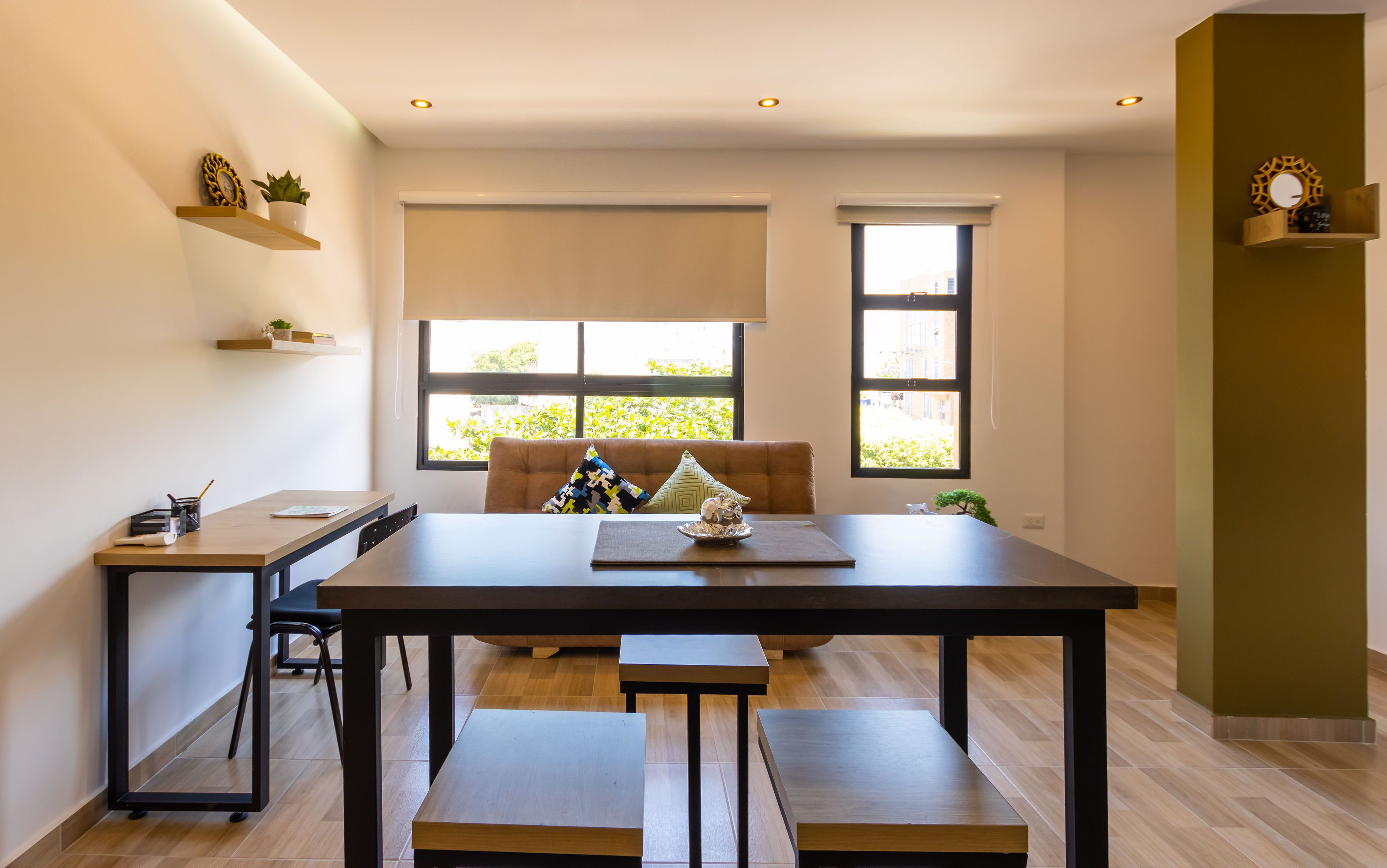 A modern dining area with a wooden table, benches, and a cushioned chair. There are two windows with blinds, a small shelf with decorative items, and a wall-mounted shelf with a potted plant.