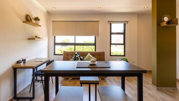 A modern dining area with a wooden table, benches, and a cushioned chair. There are two windows with blinds, a small shelf with decorative items, and a wall-mounted shelf with a potted plant.