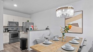 A modern kitchen with stainless steel appliances, a dining area with a wooden table set for four, and a chandelier above. - Modern 3 Bed Villa Newly Renovated Prime Location (Kissimmee)