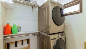 A compact laundry area with a front-loading washing machine, a dryer, and a shelf with cleaning supplies. - Modern Luxury Retreat - 2 BR/1BA - Minutes from Downtown (Rapid City)