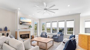 A spacious living room with a fireplace, comfortable seating, a ceiling fan, and large windows offering a view of the outdoors. - Just Shellin' - Santa Rosa Beach, Walk to Shops + Dining, Walk to Beach! (Santa Rosa Beach)