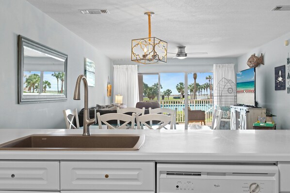 A modern kitchen with a built-in dishwasher, a dining area with white chairs, and a view of a pool and palm trees.
