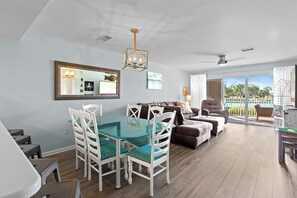 A spacious living area with a glass dining table, white chairs, a brown sofa, and a ceiling fan.