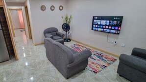Un soggiorno moderno con un divano grigio, una TV a schermo piatto montata a parete, una ventola da pavimento e un tappeto decorato.