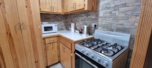 Une cuisine compacte avec des meubles en bois, un micro-ondes blanc, une cuisinière à gaz et un pare-feu en pierre.