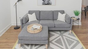 A grey sectional sofa with a matching ottoman, a floor lamp, a framed artwork of a bull, and a small side table with a plant. - Room 2-3 (Tulsa)