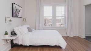 A bedroom with a bed, a nightstand, a wall-mounted lamp, a window with curtains, and a city view. - Room 2-4 Summary:<br>In (Tulsa)