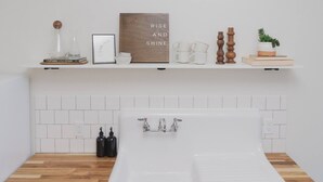 A modern bathroom with a white bathtub, wooden floor, white tiled backsplash, and shelves with decorative items and books. - Room 2-4 Summary:<br>In (Tulsa)