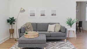 A modern living room with a grey sectional sofa, a round coffee table, and a patterned rug. There are three framed pictures on the wall, a floor lamp, and two potted plants. - Room 3-3 (Tulsa)