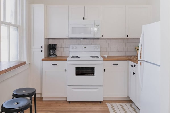 A kitchen with white cabinets, a stove, a microwave, and a refrigerator. There are two stools and a window. - Room 3-5 (Tulsa)