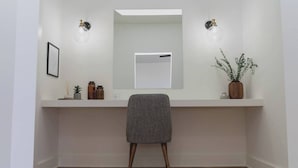 Un baño minimalista con suelo de madera, un tocador blanco con espejo, dos luces fijadas a la pared y una planta en maceta.
