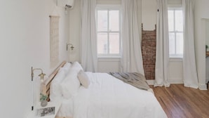 Un dormitorio con cama de madera, ropa de cama blanca, una mesita de noche con lámpara y pared de ladrillo.