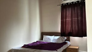 Ένα υπνοδωμάτιο με ένα κρεβάτι, ένα κομοδίνο, έναν ανεμιστήρα οροφής, μια μονάδα κλιματισμού και ένα παράθυρο με κουρτίνες.